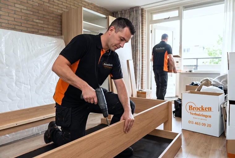Verhuizer van Brocken Verhuizingen schroeft een bedframe los, met een collega met een doos op de achtergrond
